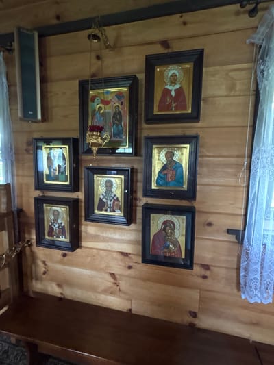 The image displays a wall paneled with light-colored wood in what appears to be a religious setting. There are seven framed religious icons hanging on the wall in a staggered arrangement. From top left to bottom right, the icons depict: two figures with halos and a golden background; a single figure in red with a halo and golden background; two figures with halos and a golden background; a male figure in blue holding a book, with a halo and golden background; a male figure with a beard and a halo, holding a book, with a golden background; a male figure in red and white vestments, holding his hands together, with a halo and golden background; and a figure in red holding a child, with halos and a golden background.

A small brass bell hangs from the ceiling by a chain, above and slightly to the left of the top-leftmost icon. To the left of the icons, near the top of the wall, is a dark gray speaker. On the far right of the image, a sheer white lace curtain is visible, partially obscuring the wooden wall. Below the icons, a dark wooden bench runs along the wall. The overall environment suggests an Orthodox Christian chapel or a similar devotional space. The location is identified as Rybaki, Poland.