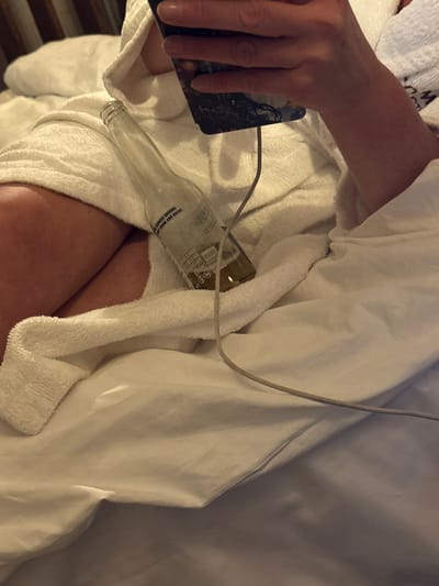 The image depicts a person in a white robe, holding a smartphone in their left hand. An empty glass beer bottle rests against their lap and the robe. A gray charging cable is connected to the phone. The scene appears to be indoors, possibly in a bed or resting area. The overall composition suggests a casual, relaxed setting.