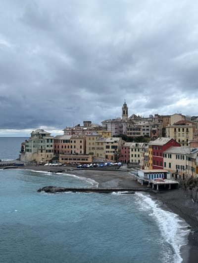 An aerial perspective captures the town of Bogliasco, Italy, under an overcast sky. The scene depicts a densely built coastal settlement with pastel-colored multi-story buildings, characteristic of Ligurian architecture, arrayed along a curved pebble beach and ascending a hillside. The Ligurian Sea, appearing turquoise near the shore and darker blue further out, displays small waves breaking on the dark grey beach. Two short, dark-rock breakwaters extend into the water.
Numerous small boats, many covered with blue or white tarpaulins, are stored on the beach in rows. A building with an orange awning, likely a restaurant, occupies a beachfront position on the left, with outdoor seating visible. To the right, a white building with blue shutters is elevated on pilings above the beach. A prominent bell tower (campanile) with a clock face and a spire rises at the highest point of the visible town, indicating a church. Scattered indistinct human figures are present on the beach and along walkways, appearing small due to the viewing distance. The overall environment is one of an active but quiet coastal town on a cloudy day.