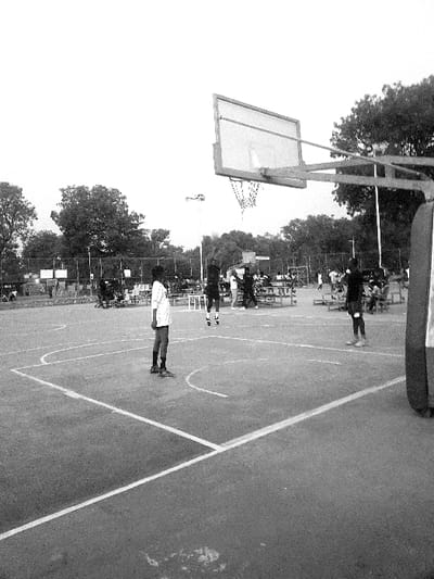 A monochrome, tilted perspective shows a basketball court with players. A hoop stands at the court's edge with the backboard catching light. Several figures, presumably players, are positioned on the court. Crisp white lines delineate the court boundaries and zones. Trees surround the open-air court, and spectators are faintly visible in the background.