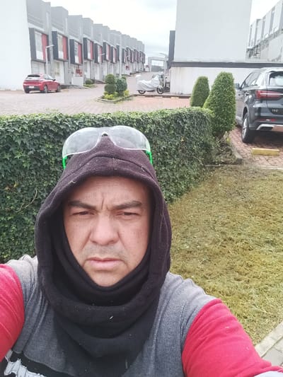 A male individual is depicted in a close-up selfie outdoors in Sangolquí, Ecuador. He wears a black fleece hood covering his head and neck, with clear safety glasses featuring green frames resting on top of the hood. His attire consists of a grey long-sleeved shirt with red sleeves. The subject maintains a neutral expression, looking directly into the camera. The background reveals a residential setting: immediately behind the subject is a dense, well-maintained green ivy hedge. To the subject's right, a patch of mowed grass is visible. Beyond the hedge, a paved area of reddish-brown interlocking bricks extends into the distance. Several conical topiary shrubs are interspersed along the paved area. Parked vehicles include a white-covered motorcycle, a dark grey SUV (Changan CS75 Plus, indicated by the "100" emblem), and a red hatchback car further in the background. A row of modern, multi-story residential units, characterized by white and dark grey facades with red architectural accents, lines the paved area. The sky is overcast.