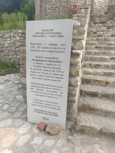 This image shows an informational plaque and the stone ruins of a monastery in Melnik, Bulgaria. The plaque, located in the foreground, is white and rectangular, featuring text in Bulgarian. The text details the history of the monastery, mentioning its construction between 1209-1211, its second name "Bogoroditsa on the Holy Mountain" and its association with the Vatopedi Monastery. It also mentions a charter from 1220, an earthquake in 1650 that caused a significant portion of the monastery to collapse, and a yearly celebration on August 31st. At the bottom of the plaque, it is labeled as a "Monument of Cultural Significance".

To the right of the plaque are stone steps, indicating the entrance or ascent to the monastery ruins. The ruins themselves are constructed of rough-hewn stones, characteristic of ancient Bulgarian architecture. Behind the plaque, partially visible, are more stone walls and some greenery, including bushes and trees, suggesting the monastery is situated in a natural, possibly hilly, landscape. Two small stones and a piece of brick are placed on the ground near the bottom of the plaque. The overall scene depicts a historical site marked for preservation and cultural tourism.