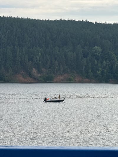 In the city of Zuevo, Russia, on a large, placid body of water, a small, dark-colored boat with a motor is navigating. The boat is occupied by two individuals. One person, wearing a light-colored top and darker bottoms, is standing at the stern, appearing to be operating the boat. Another person, in a light-colored shirt and dark shorts, is seated towards the front of the boat. A red and orange life vest is visible on the stern of the boat. In the background, a densely forested hillside rises from the water's edge, with a steep, eroded cliff face visible on the left side of the frame. The sky above is overcast with scattered clouds. In the foreground, a blue railing is partially visible.