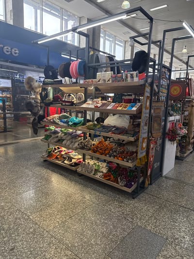 The image shows a retail display of souvenirs and traditional Kyrgyz clothing and footwear at the Bishkek Airport duty-free shop. The shop is brightly lit with fluorescent lights and has large windows offering a view of the sky. Various items are displayed on wooden shelves, including embroidered hats, slippers, textiles, and other handicrafts. A large wooden crate with a sun symbol, possibly representing the Kyrgyz flag, is visible on the right side of the display. The floor is tiled with a speckled gray pattern. The overall impression is that of a well-stocked souvenir shop catering to travelers.
