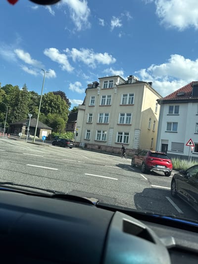 The image shows a street scene in Giessen, Germany, on a sunny day with scattered clouds. The perspective is from inside a vehicle, looking out at a row of apartment buildings and trees.

In the foreground, part of the interior of a vehicle is visible, including a dark dashboard and a portion of a blue-tinted window.

Outside the vehicle, on the street, there are several cars. A red Kia is parked on the right side of the road, with its license plate visible as "GI-CI 2503". To the left of the red car and further down the street, a black car is parked. A person on a bicycle is visible between the red car and the yellow apartment building.

The buildings appear to be residential, with multiple stories and windows. The apartment building immediately to the left of the red car is a pale yellow color, with several rows of white-framed windows. To its left, another building is visible, partially obscured by the yellow building, with a darker, red-tiled roof.

Dense green trees are visible behind the buildings and on the right side of the street, suggesting a summer or early autumn setting. A street light is present in the lower left portion of the image, extending upwards into the blue sky.

The overall impression is a typical urban street scene on a bright, clear day.