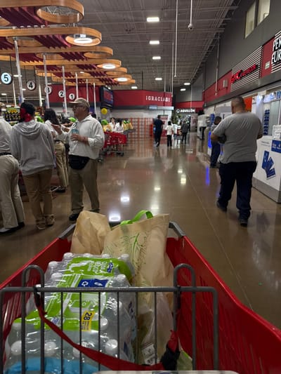 The image depicts the interior of a large retail store, identified as Abarrotes y Carnes Aguillón, located in Ciudad Santa Catarina, Mexico. The perspective is from within a red shopping cart, looking down a wide, glossy, light brown aisle that reflects overhead lighting.
In the foreground, the red shopping cart contains two multi-packs of "AGUA CUIDA MEXICO" bottled water, secured by a red strap, and two reusable tote bags: one beige with a green leaf pattern, and another plain beige.
In the midground to the left, a group of approximately six to eight individuals is gathered near checkout counters. A man in a light-colored long-sleeve shirt, khaki pants, a baseball cap, and a black fanny pack stands prominently, holding a plastic bottle and gesturing. A person in a grey hoodie with a red bow in their hair is to his left. Behind them, red and wood-paneled checkout stations are numbered 1 through 5, featuring circular light fixtures.
In the midground and background, the wide aisle extends towards the rear of the store. Several individuals are walking in the distance. A man in a grey t-shirt and dark pants walks away from the camera on the right side of the aisle. A large red wall sign displays "¡GRACIAS!" (Thank you!) in white lettering, with a smaller white sign below it reading "AGUILLÓN." Further along the right wall, a black sign with "GNC EL MEJOR" and "EURO" is partially visible. The ceiling is high, featuring exposed grey ductwork, white beams, and numerous fluorescent strip lights, along with a visible security camera.