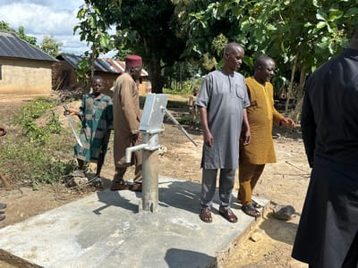 The image captures a scene in Afogo, Nigeria, focused on a newly installed water pump. Four individuals are present, with three men in the foreground gathered around the pump. The man on the far left, wearing a gray tunic and trousers, is facing away from the camera. Next to him, a man in a patterned yellow and black robe points towards the pump. To his left, a man in a brown tunic and red cap stands looking towards the pump. A young boy in a patterned green and black garment stands to the left of the pump, holding what appears to be a metal tool. The environment is rural, with a dirt ground, vegetation, and buildings with corrugated metal roofs visible in the background. The sky is cloudy. The water pump is constructed of metal and is mounted on a concrete base. The overall impression is one of a community project or an inspection of a new facility.