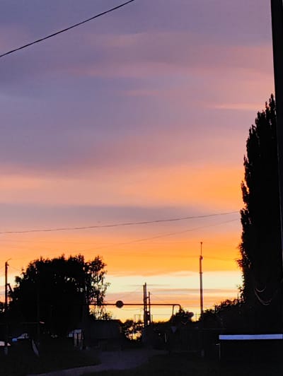 The image depicts a silhouetted view of Новоурсаево, Russia, during a sunset. The sky exhibits a gradient of colors, transitioning from a soft purple at the top to a vibrant orange and yellow near the horizon. A thin, dark power line stretches diagonally across the upper portion of the frame. In the foreground, the dark silhouettes of trees and various structures are visible, including what appear to be utility poles and wires. The overall impression is one of a rural or semi-rural landscape at dusk.