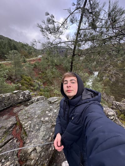 A young male, appearing to be in his late teens or early twenties, stands on a rugged, rocky outcrop in a natural, mountainous environment located in Ribeira de Pena, Portugal. He is positioned for a selfie, looking directly at the camera, wearing a dark blue hooded puffer jacket with the hood up. His right hand is visible, gripping a thin metallic cable that forms part of a railing in the lower left foreground.
The immediate surroundings feature large, grey rock formations with patches of moss and fallen pine needles. Beyond the subject, a steep valley descends, covered with dense green and reddish-brown vegetation, including numerous trees and shrubs. A winding road or path is discernible on a distant hillside, leading to a multi-arched bridge or viaduct. To the right of the subject, a stream or small waterfall is visible flowing through the valley. The sky is uniformly overcast with heavy cloud cover, suggesting cool or damp weather. A pine tree with sparse needles partially obstructs the background on the right.