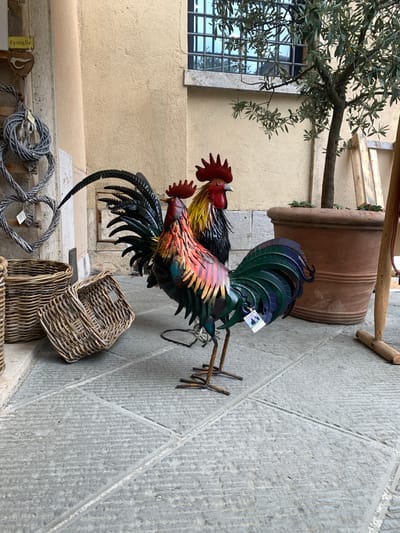 The image depicts a decorative metal rooster sculpture displayed outdoors. The rooster is vibrantly colored with iridescent blues, greens, and reds, and appears to be made of metal. It is positioned to the right of a light-colored stucco wall. To the left of the rooster, a portion of an olive tree with green leaves is visible, its branches extending over the scene. Behind the rooster and tree, a large, terracotta planter is partially visible. The ground beneath the rooster is paved with rough, grey stone tiles. In the foreground, two woven wicker baskets are stacked, and further down, more woven decorative items are arranged. A small tag is attached to the rooster's tail feathers, and a label with the word "Famiglia" is visible on a small wooden decoration in the lower left corner. The scene suggests an outdoor display, possibly at a market or shop. The lighting indicates it is daytime.