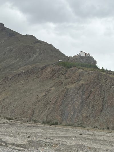 An overcast sky looms above a steep, rocky mountainside in Stongde, India. Perched precariously on the summit is a monastery, a cluster of white buildings with red accents. A line of trees, appearing as a dense, dark green band, separates the monastery from the rugged slopes below. The foreground is a dry, stony riverbed, dotted with rocks and debris, suggesting an arid landscape. Power lines are visible stretching across the mountainside, adding a modern element to the ancient scenery. The overall impression is one of isolation and grandeur, with human habitation clinging to the formidable natural landscape.