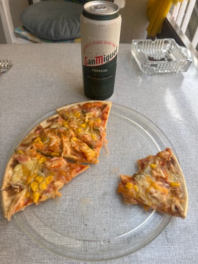 This media file shows a close-up, high-angle shot of a partially eaten pizza on a clear plastic plate, placed on a grey textured surface. Next to the plate is a can of San Miguel Especial beer. The pizza appears to be thin-crusted with melted cheese, tomato sauce, corn, and possibly some chicken or pork. One slice has been removed and is placed slightly apart from the rest. A glass ashtray is visible in the upper left corner of the frame, along with some yellow strands that could be spaghetti. The scene suggests a casual meal, possibly outdoors given the lighting. The text on the beer can is legible and reads: "CERVEZA LAGER ESPECIAL", "SURCANDO EL MUNDO DESDE 1890", "San Miguel", "ESPECIAL", "CERVEZA ESPECIAL ORIGEN ESMRA", and "DE FAMA MUNDIAL". The lighting suggests it could be daytime. The setting appears to be a table, with a hint of a striped cushion and chair visible in the lower left corner. The overall mood is relaxed and informal.