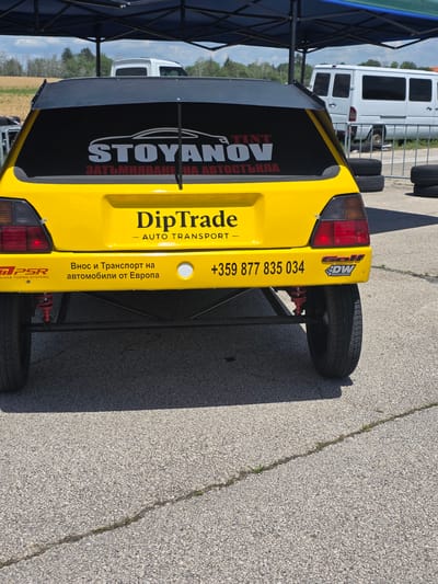 The image shows the rear of a bright yellow vehicle, possibly a custom-built or modified car, parked outdoors on an asphalt surface. The vehicle has a large black rear spoiler and a black tinted rear window. On the rear window, there is a graphic that includes the word "STOYANOV" in large white letters with a stylized car outline above it, and below that, text in Cyrillic script that translates to "AUTOMOTIVE GLASS". Below the window and above the bumper, the vehicle has several decals and text. A prominent decal reads "DipTrade - AUTO TRANSPORT". To the left of this, there is a logo with the letters "PSR" and text below it reading "PULSAR TURBO SYSTEMS". To the right of "DipTrade", there is text in Cyrillic script that translates to "Import and Transport of cars from Europe". Further to the right, there is a phone number "+359 877 835 034" and another decal with the word "GOLF" and "DW DEUTSCHWERKS".

The vehicle is partially covered by a blue and black canopy structure. In the background, a white van is visible, and beyond that, a field and trees under a cloudy sky. The asphalt surface appears to be a road or parking lot, with some cracks visible. Tires are stacked in the background.

The location is specified as the city of Akatsievo, Bulgaria. The scene appears to be a commercial or event setting, likely related to automotive services or transport. There are no people clearly visible in the image. The time of day appears to be daytime, with the sun casting shadows.