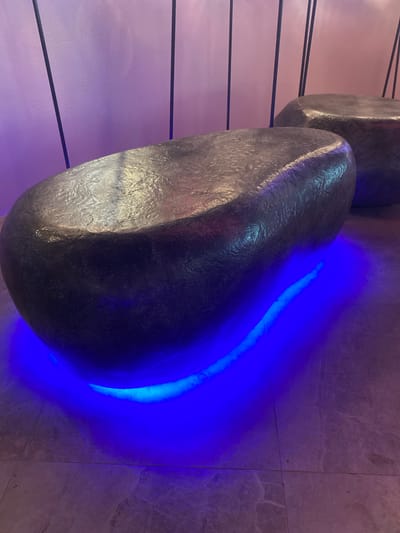 A dark, irregularly shaped bench, resembling a large, smooth stone, sits on a tiled floor. The bench is illuminated from below by bright blue LED lights, creating a vibrant glow that reflects onto the floor and the bench's underside. The bench has a textured, metallic-looking surface. In the background, a light purple wall is visible, with thin, dark vertical lines extending upwards. Another similar bench is partially visible to the right. The scene appears to be indoors, possibly in a modern public space or art installation. There are no people visible in the image. The lighting suggests it may be evening or a dimly lit environment. The flooring consists of large, light-colored tiles with a marble-like pattern. The overall aesthetic is contemporary and atmospheric. The location is Berdsk, Russia.