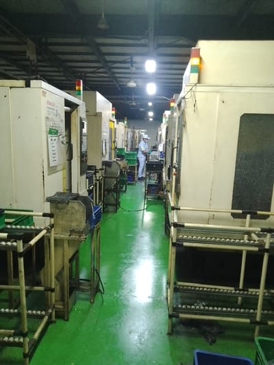 The image shows a factory setting with rows of large industrial machines. The floor is a bright green, and the ceiling is high with exposed beams and lighting fixtures. In the background, a person in light blue work clothes and a white hat is operating one of the machines, wearing a face mask and gloves. They appear to be engaged in a manufacturing or assembly process.

Several machines have prominent tower lights on top, with red, amber, and green indicators, likely signaling operational status. Some of these lights are illuminated. There are also various bins, some green and some blue, placed around the machinery. In the foreground, there are racks with what appear to be small spherical components, possibly for manufacturing.

On the side of one of the machines, there is visible text, including "FANUC ROBODRILL" and "INSTRUCTION KERJA". The overall impression is a functional, active industrial environment. The lighting suggests it is daytime inside a well-lit facility. The presence of safety equipment on the worker and the organized nature of the factory indicate a focus on production and order. The location is identified as Ciantra, Indonesia.