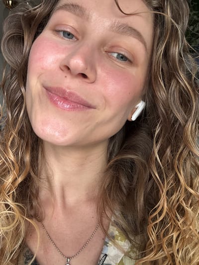 A close-up portrait of a woman with curly, light brown and blonde hair. She has light skin, blue eyes, and is wearing a white earbud in her left ear. A silver necklace is visible on her neck. There is no evidence to suggest the location is in Ketoshi, Russia. The environment is not distinctly identifiable, suggesting the focus is on the individual.