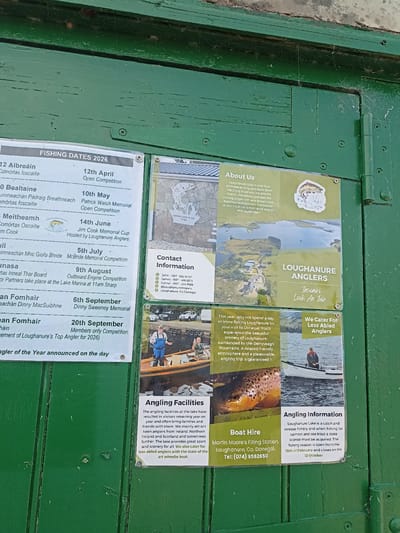 This is a photograph of a wooden door painted green, likely on the exterior of a building in Loughanure, Ireland. The door is weathered and has visible spiderwebs. Attached to the door are several informational posters related to fishing.

The posters detail fishing dates for 2026, including various competitions and memorial events such as the "Patrick Walsh Memorial Open Competition" and the "Dinny Sweeney Memorial." One poster provides "Contact Information" with phone numbers and social media handles for "Loughanure Anglers."

Another poster, titled "About Us," describes Loughanure Lake as a fishery offering brown trout, sea trout, and salmon, and notes that the salmon and sea trout run occurs from early May to mid-September. There is an illustration of a leaping trout.

A section on "Angling Facilities" highlights the lake's appeal to both local and international anglers, mentioning that it caters to less-abled anglers with a specialized "wheelie boat."

A "Boat Hire" poster advertises services from "Martin Moore's Filling Station."

Finally, an "Angling Information" poster explains that Loughanure Lake is a catch-and-release fishery, requiring a state license for salmon and sea trout fishing, with the season running from February 15th to October 12th. The overall impression is that of an advertisement for local fishing opportunities.