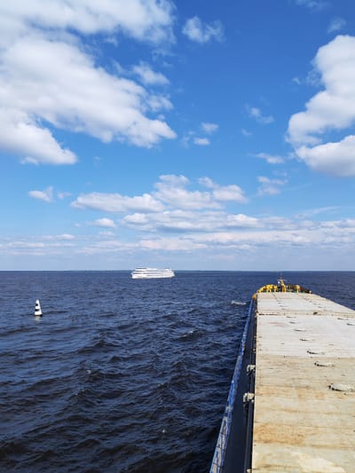 The image depicts a wide, dark blue body of water under a bright blue sky with scattered white clouds. The perspective is from the deck of a vessel, looking out towards the horizon. In the distance, a white passenger ship with multiple decks is visible, cruising across the water. Closer to the foreground on the left, a black and white striped buoy stands upright in the water. The right side of the image shows the deck of the vessel from which the photo is taken, characterized by a large, flat, grey surface with visible seams and circular latches. The water is choppy with moderate waves. The scene is set in Veregonets, Russia, on a clear day.