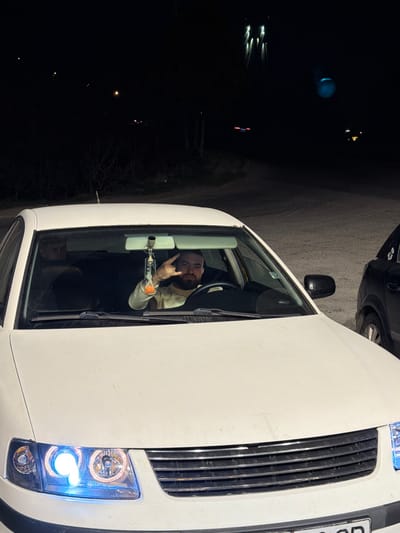 A white vehicle, consistent with a Volkswagen Passat (B5/B5.5 generation), is positioned at night in what appears to be a parking area or unlit roadside in Agigea, Romania. The vehicle's modified headlights, featuring an illuminated blue ring on the left side, are active. The driver, a bearded male wearing a light-colored garment, is visible through the windshield and is performing a 'rock on' hand gesture with his left hand. An hourglass-shaped air freshener or decorative item hangs from the rearview mirror. To the right of the white vehicle, the front fender and wheel arch of a dark-colored vehicle are partially visible. The background is dark, with distant faint lights suggesting streetlights or other structures, and sparse, possibly bare, vegetation. The ground surface appears to be asphalt or compacted dirt.