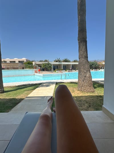 The image depicts a person lying on a lounge chair, with their legs extending into the foreground, a bright blue swimming pool in the middle ground, and resort buildings in the background. A palm tree trunk is on the right side of the frame, partially obscuring the view. The sky is clear and blue. The person is wearing a dark nail polish on their toes. The pool appears to be in the city of Tavronitis, Greece, with several people visible in and around the water. One person in red shorts stands by the pool, and others are swimming. The resort buildings are modern, with white walls and flat roofs. The overall impression is one of a relaxing vacation scene.