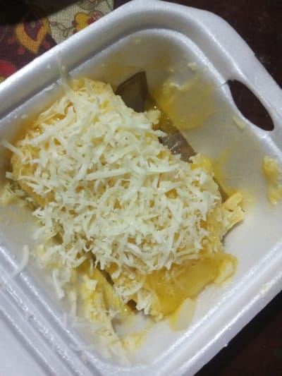 The scene depicts a white styrofoam container holding a food item. The container is partially filled with a yellow, semi-liquid base, heavily topped with finely shredded white cheese. Below the yellow base, darker, irregular shapes, possibly pieces of a solid food component, are visible. The yellow substance exhibits a glossy or oily texture with small bubbles. The container features an oval-shaped indentation or void on its upper right rim. The background on the upper left shows a blurred, patterned fabric with red, yellow, and brown designs, while the remaining background is dark and indistinct. No individuals or discernible actions are present. The specified location, "Pandaria, India," is not verifiable as Pandaria is a fictional region.