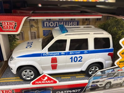 This is a close-up, eye-level shot of a toy police car, a white SUV, displayed in its packaging. The toy car has blue and white detailing, including the word "ПОЛИЦИЯ" (Police) and the number "102" on its side. It also features a blue light bar on the roof. The packaging depicts a building facade with the word "ПОЛИЦИЯ" above an entrance. A red triangular warning sign with an arrow and the word "НАЖМИ!" (Press!) is visible in the foreground. The background shows shelves in what appears to be a retail store. The overall impression is of a product display for a children's toy. The image does not contain any information or visual cues that would allow for the identification of the city of Kuzmolovsky, Russia.