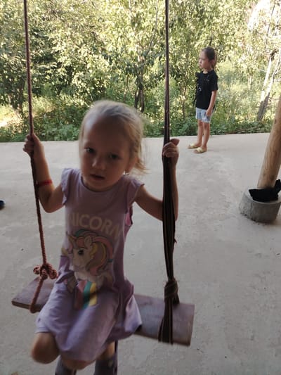 In Shovot, Uzbekistan, two young girls are present outdoors on a concrete patio. The girl in the foreground, seated on a wooden swing, is wearing a purple sleeveless dress with a unicorn graphic and the word "ICORN" visible. She has blonde hair and appears to be looking towards the camera. The girl in the background is standing and wearing a black t-shirt and denim shorts. She is looking to her left. The area behind them is covered with dense green foliage. A concrete pillar with a black object on top is situated on the right side of the image.