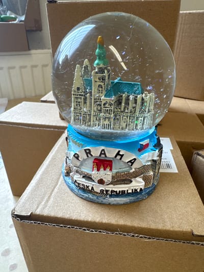 An indoor scene in Neratovice, Czechia, displays a single snow globe positioned centrally on a brown corrugated cardboard box. The snow globe features a clear spherical dome filled with liquid and numerous iridescent flakes, surrounding a miniature architectural model. This model depicts a prominent, light-colored building complex with multiple spires and a distinctive teal-colored roof, resembling a cathedral.

The snow globe rests on a blue resin base adorned with relief carvings. The word "PRAHA" is visible in white capital letters along a raised arc, with "ČESKÁ REPUBLIKA" inscribed below it. Other details on the base include a stylized rendering of a bridge, possibly depicting Charles Bridge, with a tower-like structure, and a small boat on sculpted water. A miniature depiction of the flag of the Czech Republic is also present on the base.

The snow globe is surrounded by multiple brown corrugated cardboard boxes, stacked and arranged in the background, suggesting a storage or packing environment. A white radiator is partially visible in the far left background. A white adhesive label, with text including "01 2026 VSSV" and other alphanumeric characters, is affixed to a box in the mid-right background. No individuals or active interactions are depicted.