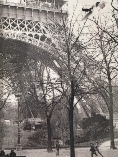 This black and white photograph captures a view of the Eiffel Tower seen through a dense network of bare tree branches. The image is rotated 90 degrees counter-clockwise. The intricate lattice structure of the tower is visible on the left, with its metal framework forming a geometric pattern. The trees, devoid of leaves, create a stark, almost skeletal appearance against a bright sky.
In the background, on the right side of the frame, a path runs alongside a fence. A few figures are visible on this path, appearing as silhouettes against the lighter ground. One individual, closer to the foreground, is bent over, possibly in motion. Further along, two figures stand upright. The scene suggests a park-like setting, likely during a time of year when trees have shed their foliage, possibly autumn or winter. The overall mood is somewhat melancholic and stark due to the bare branches and the black and white rendering.
Some text is visible on the side of the Eiffel Tower structure, including what appears to be "SOUTENE" and "BICHAT". The rotation of the image makes it difficult to read any further text or discern specific details about the time of day or weather beyond the general impression of a bright, possibly overcast, day. The location is identified as Molenbeek-Saint-Jean - Sint-Jans-Molenbeek, Belgium, but the Eiffel Tower is famously located in Paris, France, suggesting a possible misidentification of the landmark or an artistic interpretation.