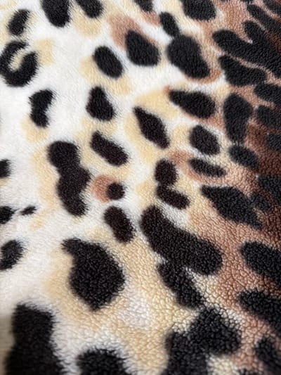 The image is a close-up, detailed view of a leopard print fabric. The texture of the fabric is soft and plush, resembling fleece or faux fur. The print consists of irregular black spots scattered across a background of creamy white and light tan or beige patches, mimicking the natural pattern of a leopard's coat. Some of the black spots are solid, while others appear to have a lighter, brownish-pink center, adding depth and realism to the pattern. The lighting highlights the fluffy texture of the material, with shadows and highlights emphasizing the raised nap of the fabric. There is no indication of a specific location within the city of Vilshany, Ukraine, as the image is solely focused on the fabric's pattern and texture.