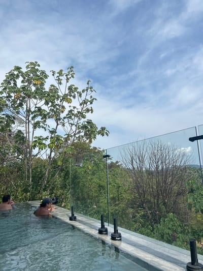 The image shows a swimming pool with a glass railing and a view of trees and a blue sky with white clouds in the background. There are two people in the pool, one sitting on the edge and another swimming. The location is at coordinates 9.647, -85.166, which is in the city of Santa Fe, Costa Rica. The scene is likely at a hotel or resort, with the swimming pool and railing suggesting an upscale environment. The individuals in the pool are likely relaxing or enjoying the view. The clear water and surrounding vegetation add to the feeling of tranquility and serenity. The image captures a moment of leisure and relaxation.