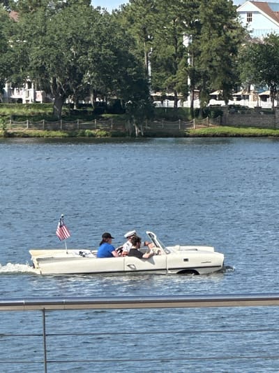 The scene presents a boat navigating a body of water, likely a lake or canal, in Lake Buena Vista, Florida, United States. The boat is light-colored and bears a small American flag. Several individuals are visible on board, including one wearing a blue shirt. The boat is leaving a wake. A building and trees are on the left side of the image. There is a structure on the right side of the image.