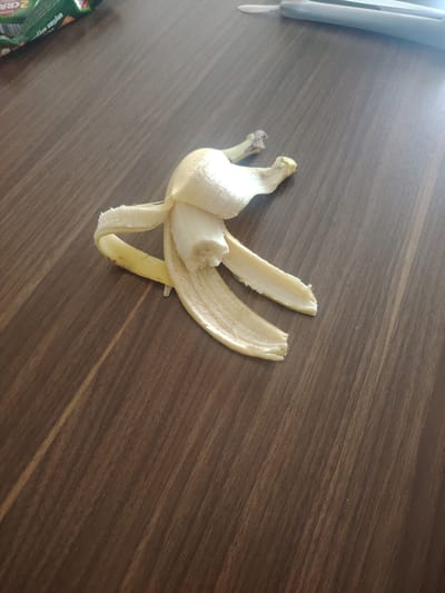 The image shows a banana peel lying on a wooden surface. The peel is mostly yellow with some brown spots. It appears to be partially opened. The wooden surface has a visible grain pattern. In the upper left corner, a green package with writing is partially visible, and in the upper right corner, a gray object, possibly a ruler or a pen, is visible. The scene is indoors, on a table. The image does not contain any information that can be used to determine its location in the city of Hovsan, Azerbaijan.