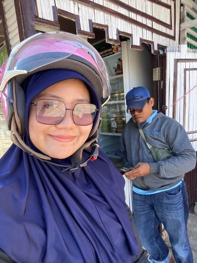 In the city of Sungai Paring, Indonesia, a woman wearing a purple hijab and a pink helmet is taking a selfie. She is also wearing large square-framed glasses. Behind her, a man wearing a blue baseball cap, a gray hoodie with a light blue undershirt, and blue jeans is looking at his phone. He has a small green bag slung across his chest. They appear to be standing in front of a small storefront with a white wooden exterior and a door that is slightly ajar, revealing a refrigerator stocked with items inside. The overall scene suggests an outdoor marketplace or a small retail establishment.