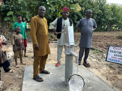 The image depicts an outdoor scene in Afogo, Nigeria, where a group of individuals, including several men and two children, are gathered around a manual water pump. The pump is operational, with water flowing into a clear plastic bucket positioned beneath its spout. The ground surrounding the pump is a mixture of dry earth and some tilled soil, suggesting an agricultural or rural setting. Lush green foliage, likely trees and bushes, forms a backdrop to the scene.
In the foreground, a man dressed in a distinctive yellow and black patterned traditional outfit stands to the left, facing the camera. To his right, another man, wearing a light-colored outfit and a red cap, is operating the pump handle. Further to the right, a third man in a grey traditional outfit stands with his hands on his hips, looking away from the camera.
Two children are also present. One, a young boy in a faded t-shirt and shorts, stands to the left of the man in the yellow and black outfit. Another child, partially visible on the far left, is dressed in a patterned shirt and dark trousers.
A white sign with black lettering is positioned on the right side of the frame. Although partially obscured, it appears to be a donation acknowledgment, mentioning "DON" and "NORTHERN ASSOCI" and "FOUNTAIN OF LI". The overall atmosphere suggests a community event or the inauguration of a water source.