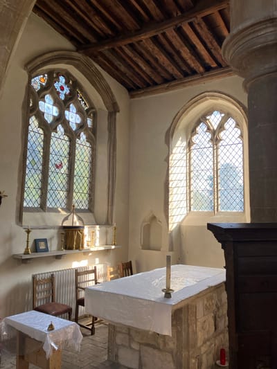 The image displays the interior of a stone church or chapel, located within the geographical area of Mid Suffolk, United Kingdom. No specific identifying architectural elements of a particular edifice are present.
The scene features two distinct arched, multi-pane windows with stone tracery. The larger window on the left exhibits clear diamond-patterned leaded glass, with decorative stained glass elements in its upper portion, including red and blue floral designs, and a small red bird or leaf motif. The smaller window on the right contains clear diamond-patterned leaded glass. Direct sunlight streams through both windows, casting prominent grid-like shadows onto the interior walls and floor.
The room's walls are light-colored, possibly plastered, and a ceiling constructed from exposed dark wooden beams is visible overhead. A large, stone-built altar or table in the foreground on the right is covered with a translucent white plastic sheet. A single unlit white taper candle, held in a brass candlestick, is centered on this altar. A small red cylindrical object rests on the floor at the altar's base, towards the right.
To the left, a smaller wooden table is also draped with a similar white translucent plastic sheet. A small brass bell with a wooden handle is positioned on this table. Beneath the large left window, a stone shelf is affixed to the wall. Items arranged on this shelf include two brass candlesticks, each holding an unlit white candle, a small framed picture with a blue mat, a gold-fabric-wrapped cylindrical vessel, and a clear glass vessel. A white radiator is installed directly below this shelf.
Three wooden chairs are visible in the midground: two with wooden frames and woven seats, and one with a dark frame and a leather-like seat. An empty, arched stone niche is set into the right wall. A large, fluted stone pillar is partially visible on the far right edge of the frame. No individuals are present in the scene.