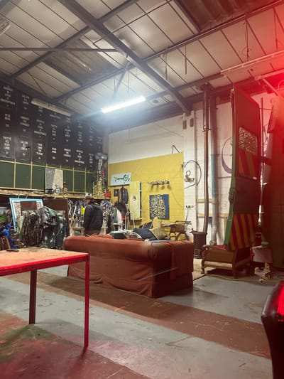 The image depicts an indoor scene, likely a large warehouse, workshop, or a repurposed retail space, shown rotated 90 degrees counter-clockwise.
The setting features high ceilings with exposed metal beams and trusses, indicative of an industrial building. Fluorescent strip lights illuminate the space. The floor is concrete, with distinct sections painted in gray and a reddish-brown color. Walls are painted in varying tones, including a bright yellow section in the middle-ground and white/off-white elsewhere. Exposed pipes and electrical conduits run along the walls and ceiling.
In the upper portion of the image, a large, dark green vehicle or piece of industrial machinery is visible, partially obscured. It features distinct red and yellow diagonal striped markings on its side. Near this vehicle, a woven wicker chair is present.
Towards the right side of the image, a substantial brown upholstered sofa or sectional couch occupies a significant portion of the floor space. In the immediate foreground, a red metal table or workbench is partially visible, extending from the bottom right corner.
Against the yellow wall, several artworks or decorative pieces are displayed, including a framed mosaic-like piece with many small colorful components and another featuring abstract green shapes. Beneath these, numerous items of clothing or textiles are arranged on racks or shelves, contributing to a busy, potentially retail-oriented atmosphere.
On the left side, a large section of wall is covered with black panels or posters, each bearing white text. A person wearing a dark jacket is visible among these displays, seemingly browsing or organizing items.
The space appears somewhat cluttered and functional, suggesting it might be a thrift store, a vintage shop, an artist's studio, or a storage area. The lighting is artificial, and a red lens flare or light source glare is noticeable in the upper left corner of the original image.
Visible text on the black panels includes phrases such as:
- "BUSINESS AS USUAL"
- "CALLING ALL EARTH"
- "UNACCEPTABLE"
- "BENT LIVE"
- "READ MORE"
- "EMBRACE THE STRUGGLE"
- "THEY'RE ALL BENT LOVE"
- "THEN CAME THE PLAGUE"