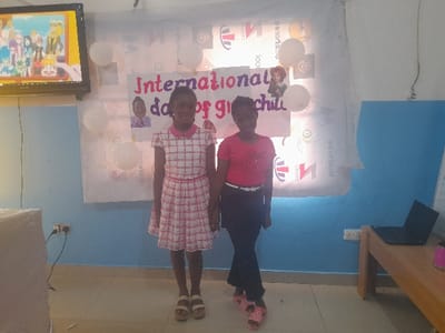 The image depicts an indoor scene in Ajah, Nigeria, featuring two young females standing in front of a celebratory backdrop. The wall behind them is painted light blue at the bottom and white at the top. A large white banner is affixed to the upper wall, displaying the red text "International day of girl child" and several logos, including "Rotary." Six white balloons are attached to the banner, along with two small portrait images of young girls.
On the left, a flat-screen television is mounted on the wall, displaying an animated scene with multiple characters. Below the television, a white object covered by a white cloth is partially visible. An electrical outlet with connected wires is present on the blue wall section to the left.
On the right side of the image, a light brown wooden table is positioned against the wall. A black laptop, open, is on the table, along with a pink cylindrical object. An electrical outlet is visible on the blue wall beneath the table.
Two young females are the central subjects. The female on the left has dark hair, wears a white and pink plaid short-sleeved dress with a pink collar and belt, and light-colored platform sandals. She stands facing forward with a slight smile. The female on the right has dark hair, wears a pink short-sleeved t-shirt with a design, dark trousers, and pink patterned slides. She stands facing forward with her legs crossed and a slight smile.