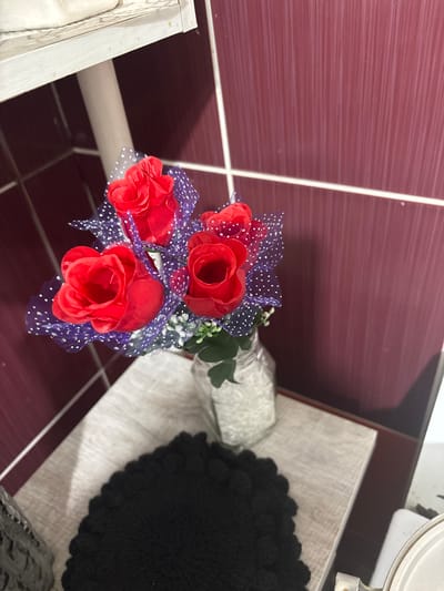 A bouquet of artificial red roses with purple polka-dot mesh ribbon is displayed in a glass jar. The roses appear to be made of fabric and have a slightly crumpled texture. They are tied together with the mesh ribbon, creating a decorative effect. The jar containing the roses is filled with white, shredded material that could be paper or a similar filler.
The background consists of a tiled wall with a dark reddish-purple hue. The tiles are laid horizontally, and the grout lines are visible as thin white stripes. To the right of the bouquet, a textured, light-colored surface, possibly a table or shelf, is visible. On this surface, a black, round object with a textured, possibly crocheted surface, is placed.
The lighting in the image suggests an indoor setting, possibly with artificial light. There are no indications of the time of day or weather conditions. The overall scene is still and decorative, with no people or active events taking place. The focus is on the artificial flower arrangement. No visible text is present in the image. The location context provided is Княгинино, Russia.