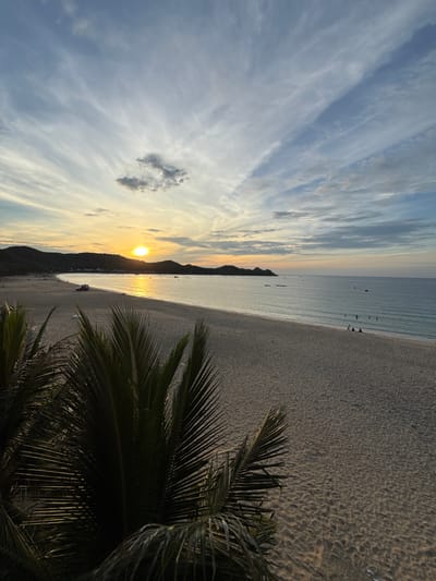 This image captures a serene sunset over Xuan Dai Bay in Song Cau, Vietnam. The sky is a canvas of soft blues and grays, streaked with wispy white clouds, with the sun casting a warm golden glow as it dips towards the horizon. The calm, turquoise waters of the bay gently lap against a wide expanse of sandy beach. In the distance, a line of dark, rolling hills forms the coastline, dotted with small buildings. A few small boats are visible on the water. On the beach, a red vehicle is parked near the water's edge, and a few individuals are scattered across the sand, some wading in the shallow water. In the foreground, the fronds of a palm tree frame the scene, adding a touch of tropical lushness. The overall atmosphere is one of tranquility and natural beauty.