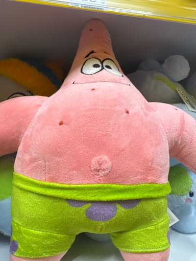 A close-up, eye-level shot features a stuffed toy of Patrick Star from SpongeBob SquarePants, positioned on a shelf in a retail environment, likely a toy store or department store in Ivanivka, Ukraine. The plush toy is predominantly pink, with a star-shaped body, and wears green shorts with purple polka dots. The toy's face is visible, displaying a cheerful expression with large white eyes and a wide smile. Red dots are scattered across its body, mimicking freckles. The background is blurred, showing other plush toys and the shelving units. A yellow shelf liner is visible at the top of the image, suggesting it is part of the store's display. The lighting appears to be artificial, typical of an indoor retail setting.