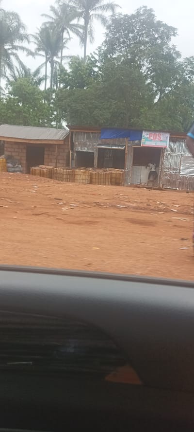 The image shows a street scene in what appears to be Oto, Nigeria. The scene is dominated by two small, adjacent buildings made of corrugated metal and unfinished brick. One building has a sign that reads "POS" (Point of Sale), and a person is standing inside the building. In front of both buildings, there are several containers, likely filled with liquid. The ground is red dirt, and in the background, there are palm trees and other vegetation. The image appears to have been taken from inside a vehicle, as part of a car window is visible in the foreground. The overall impression is that this is a rural or developing area of Oto.