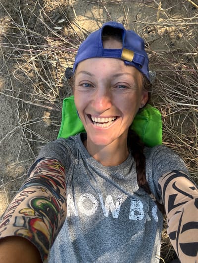 A person is lying on dry grass and dirt, looking up at the camera. They are wearing a blue baseball cap and a grey t-shirt. The person has dark brown hair braided and resting on their shoulder. They are smiling broadly, showing their teeth, and have blue eyes. Colorful tattoos are visible on their arms and legs, with one on the leg appearing to have a black abstract pattern and another on the arm featuring bright colors, possibly depicting eyes and flames. A bright green object, possibly a small pillow or bag, is under the person's head. The ground is covered in dry, straw-like grass and some patchy dirt. The lighting suggests it is daytime, with sunlight casting shadows and highlighting the textures of the ground and grass. The overall mood appears cheerful and relaxed. Visible text on the t-shirt is partially obscured but appears to say "WORK" in large, distressed white letters.