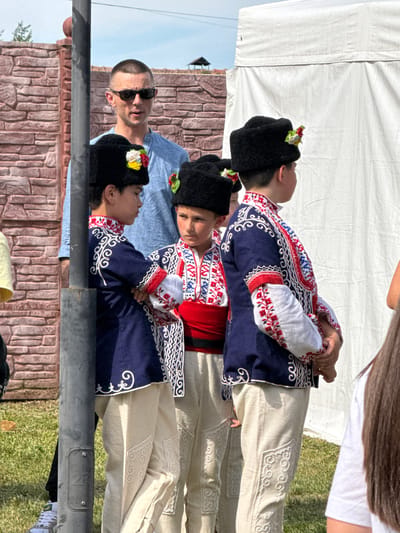 In Pordim, Bulgaria, a man wearing sunglasses and a blue shirt stands behind three boys dressed in traditional Bulgarian attire. The boys are wearing dark, fur-trimmed hats adorned with colorful flowers, embroidered blue vests with red and white trim, red sashes, and off-white pants with intricate white embroidery. They are standing in a line, facing right, and appear to be participating in a cultural event. Behind them, a rustic brick wall forms the backdrop, and a white tent is visible on the right side of the image. The ground is covered in green grass. The man appears to be supervising the boys.