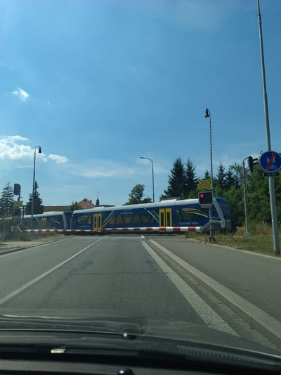 The image captures a street-level view from a vehicle looking towards a railway crossing in Horažďovice, Czechia. A blue and white train, bearing the text "PLZEŇSKÝ KRAJ," is stopped at the crossing, its barrier down. Red signal lights are illuminated, indicating the train's presence. Adjacent to the crossing, a sign with a red circle and a diagonal line over a bicycle and a pedestrian signifies a prohibition for these modes of transport. Trees and vegetation line the background, with a clear blue sky overhead. The road surface shows lane markings and a noticeable dark stain.