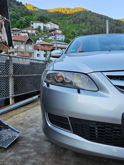 The image shows the front of a silver Mazda 3 parked on a metal surface, possibly a trailer or flatbed truck. In the background is a hillside town, identifiable as Erma Reka, Bulgaria, based on the characteristic architecture and topography. The houses are primarily two or three stories, with red tile roofs. The hillside is covered in trees, with some signs of spring foliage. The Mazda has its hood slightly open, revealing the grill. The front bumper and headlight are in focus. The metal surface beneath the car appears worn and rusty. A chain-link fence is visible in the foreground. No people are visible in the scene.