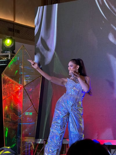 A woman in a shimmering silver jumpsuit is performing on a stage in front of a large LED screen. She has her hair pulled back in a high ponytail and is wearing large hoop earrings. She is leaning forward, with her right arm extended and pointing downwards towards a geometric, multi-faceted mirrored structure on the stage. The LED screen behind her displays abstract, wavy patterns in shades of grey, white, and purple. The stage is illuminated with colorful lights, including red, blue, and green, which reflect off the mirrored structure. A green spotlight is visible in the lower left corner of the image, and a black object, possibly a camera or speaker, is partially visible in the upper right corner. The woman appears focused and energetic, likely engaged in a dance or musical performance. The overall atmosphere is dynamic and theatrical, suggesting an indoor or evening event.