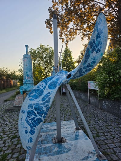 The image depicts a large, stylized sculpture resembling a propeller, painted with a blue and white abstract pattern resembling water or ice. The sculpture is mounted on a metal frame and appears to be outdoors. The setting includes a cobblestone pathway with grass growing between the stones, trees with golden foliage, and greenery. In the background, there's a clear sky, suggesting a pleasant day. A small sign with red and white text is visible on a pole amidst the bushes. There's also graffiti on a concrete structure to the right of the sculpture.

The sculpture itself has visible text on one of its blades: "KSB" logo and "HALLE 1200 JAHRE". This indicates the sculpture is related to the company KSB and commemorates the 1200th anniversary of Halle, Germany. The time of day appears to be late afternoon or early evening, as indicated by the warm light illuminating the trees and the sky. The overall mood is calm and artistic, with the sculpture being the central focus against the natural and urban elements of the environment.