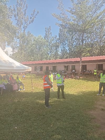 An outdoor gathering is depicted on a bright, sunny day in a grassy area within Idakho South ward, Kenya. In the immediate foreground, a man wearing an orange reflective safety vest stands facing right, his back partially to the camera. To his right, a second man in a lime green reflective safety vest faces towards the left, appearing to speak into a small, dark object possibly a microphone.
To the left, a large white canopy tent is partially visible, under which several individuals are seated on chairs. Some of these individuals are also wearing lime green reflective safety vests. The ground is covered in short, green grass, with some patches of shadow cast across it.
In the mid-ground, a single-story building with light-colored walls and a red corrugated iron roof is visible. The building has multiple window and door openings. Scattered in front of and around this building are numerous other individuals, some seated on what appear to be benches or on the ground. Several of these individuals also wear lime green reflective safety vests.
The background features numerous tall, deciduous trees with dense green foliage against a clear, bright blue sky.