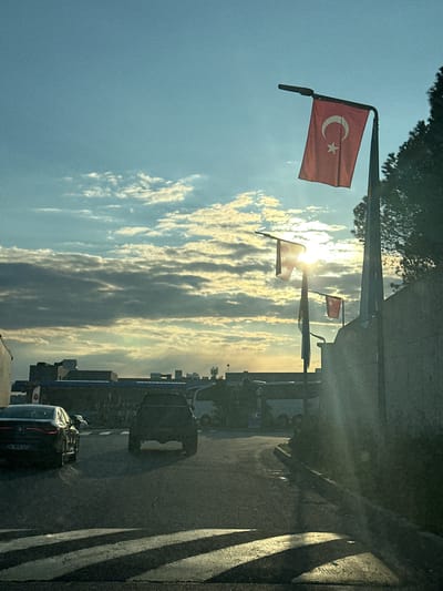 The image shows a street scene in Gaziosmanpaşa, Turkey, with Turkish flags hanging from poles along the roadside. The sky is visible, with bright sunlight filtering through the clouds. Several cars are on the street, including a dark-colored car in the foreground. The right side of the frame shows a sidewalk with pedestrian markings. Buildings and other infrastructure are visible in the background.